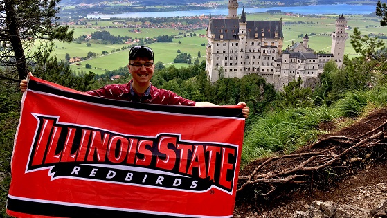 Student holding an Illinois State Redbirds flag in front of Neuschwanstein Castle in Germany