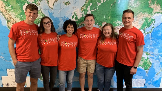 Six study abroad ambassadors wearing matching Roaming Redbirds t-shirts pose in front of a world map